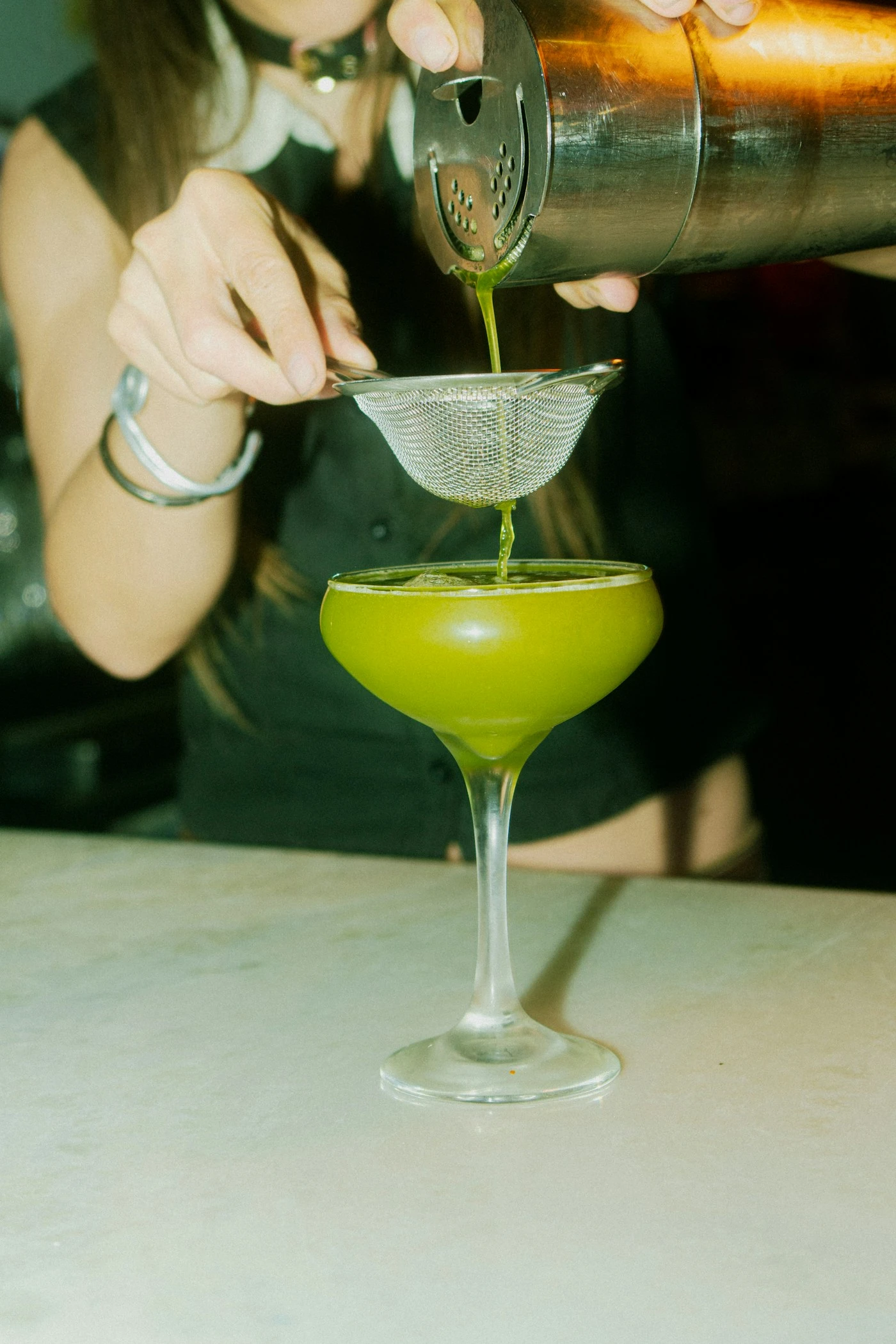 Bartender pouring a bright green Grasshopper-style cocktail into a chilled coupe
