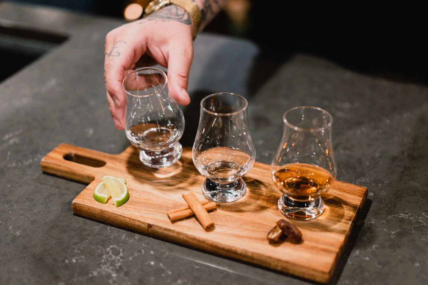 Homemade Cinnamon Whiskey: A hand with tattoos holds a whiskey glass on a tray with cinnamon sticks and lime. Perfect for lifes