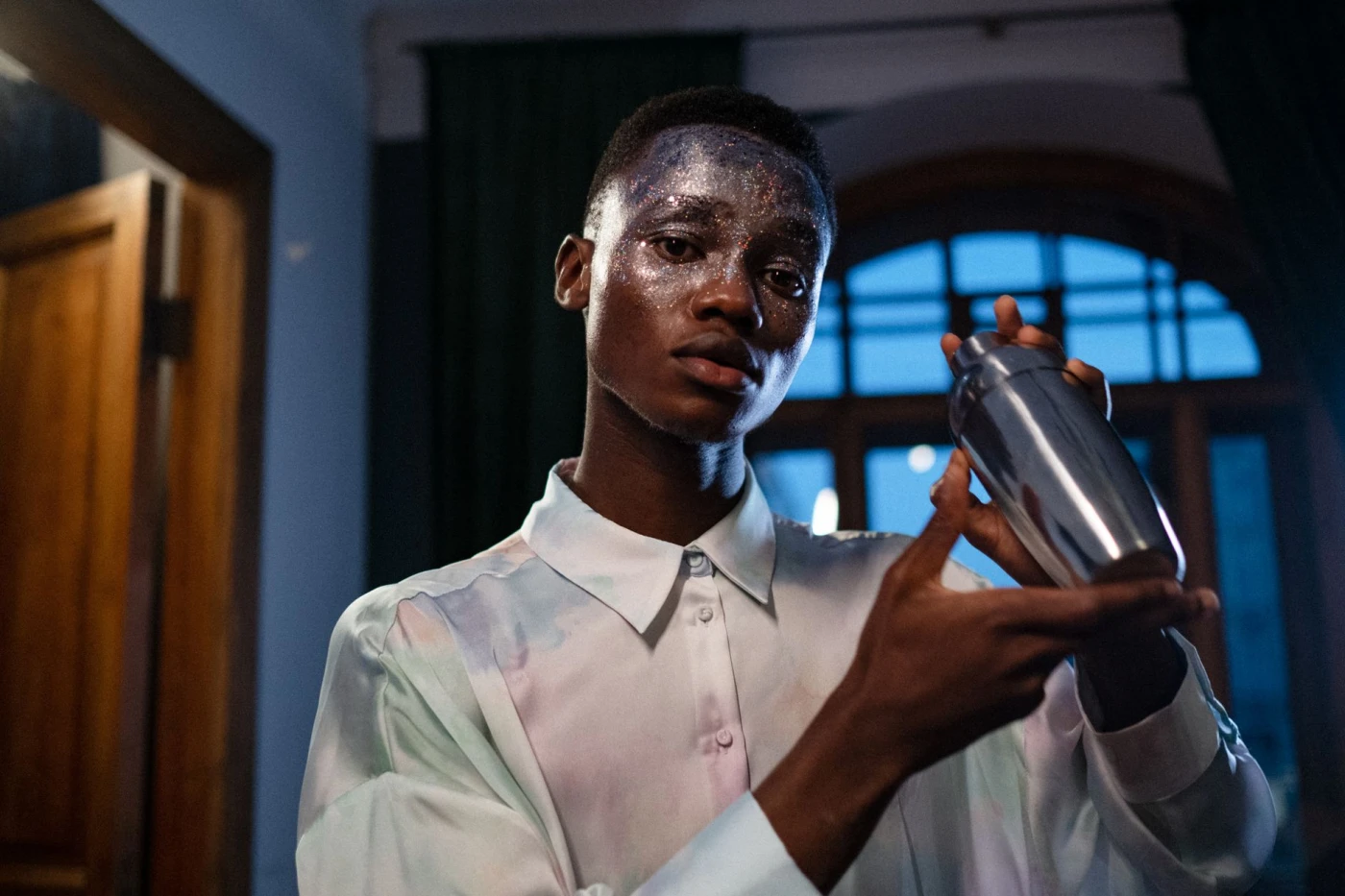 Stylish young man with glitter makeup holding a cocktail shaker in an indoor setting.