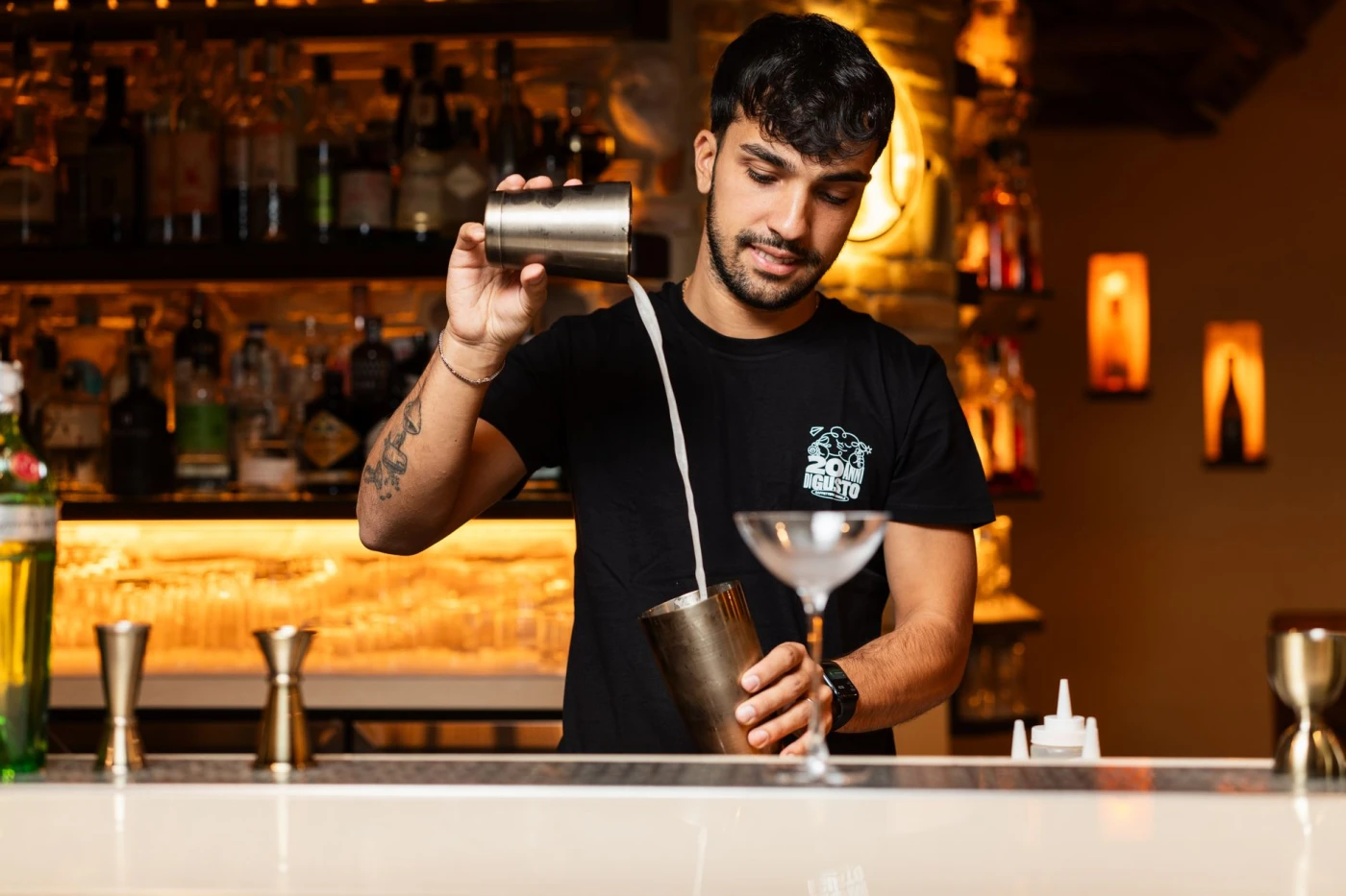 A bartender expertly mixes a cocktail behind a bar, pouring liquid into a shaker with a vibrant background.