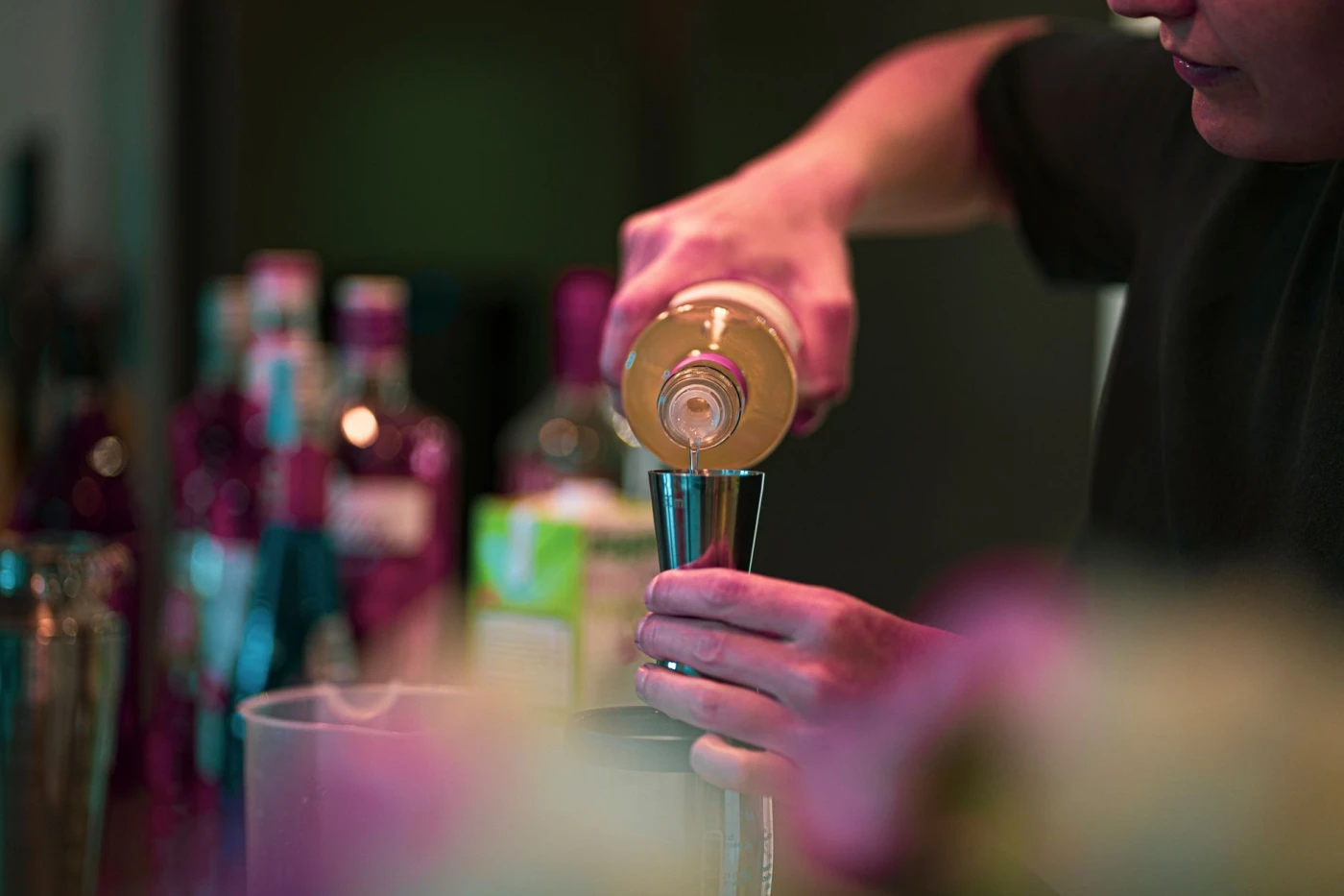 A bartender pours a drink into a jigger at a stylish bar with bottles in the background.
